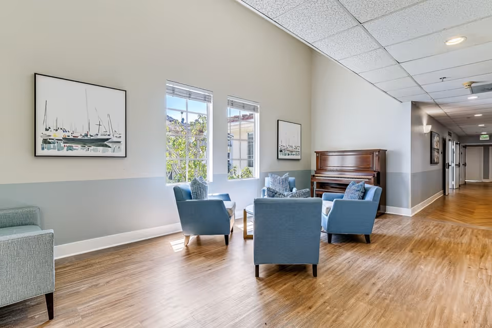 A bright and spacious senior living facility common area with four blue armchairs arranged around a small round table near two large windows. A wooden piano is positioned against the wall, and two framed paintings of sailboats hang on the walls. The floor is wood, and the ceiling has recessed lighting. A hallway extends to the right.