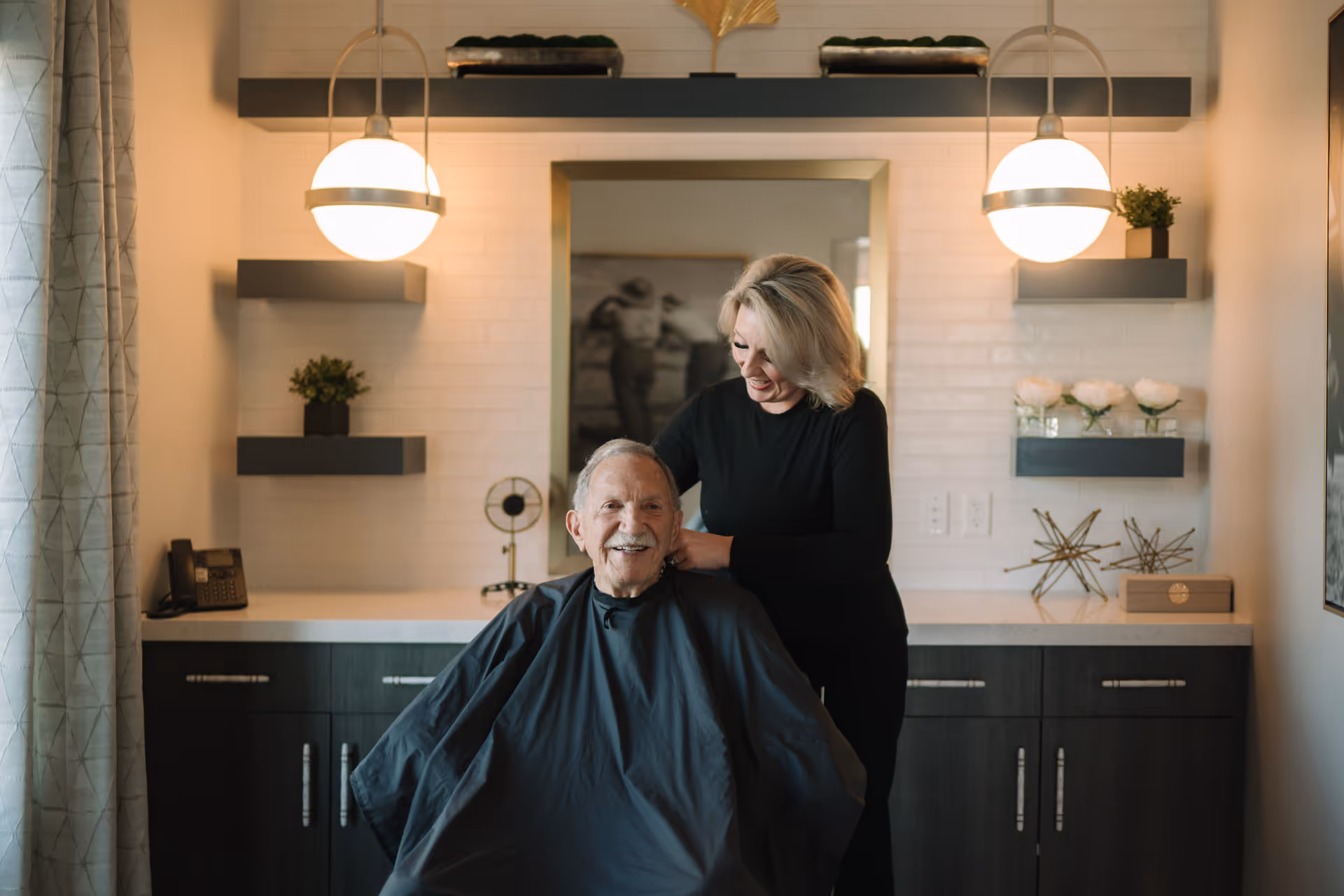 An elderly man sitting in a chair with a black haircut cape on, smiling while a woman standing behind him styles his hair in a well-lit room with modern decor including shelves with plants and decorative items, a large mirror, and two hanging globe lights.