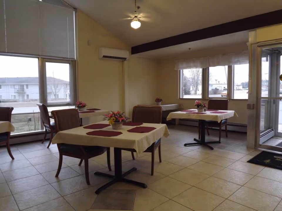 A small dining area with four tables covered with white tablecloths and red placemats. Each table has a small floral centerpiece. The room has large windows with blinds and a glass door leading outside. There are several brown cushioned chairs around the tables, and the floor is tiled.