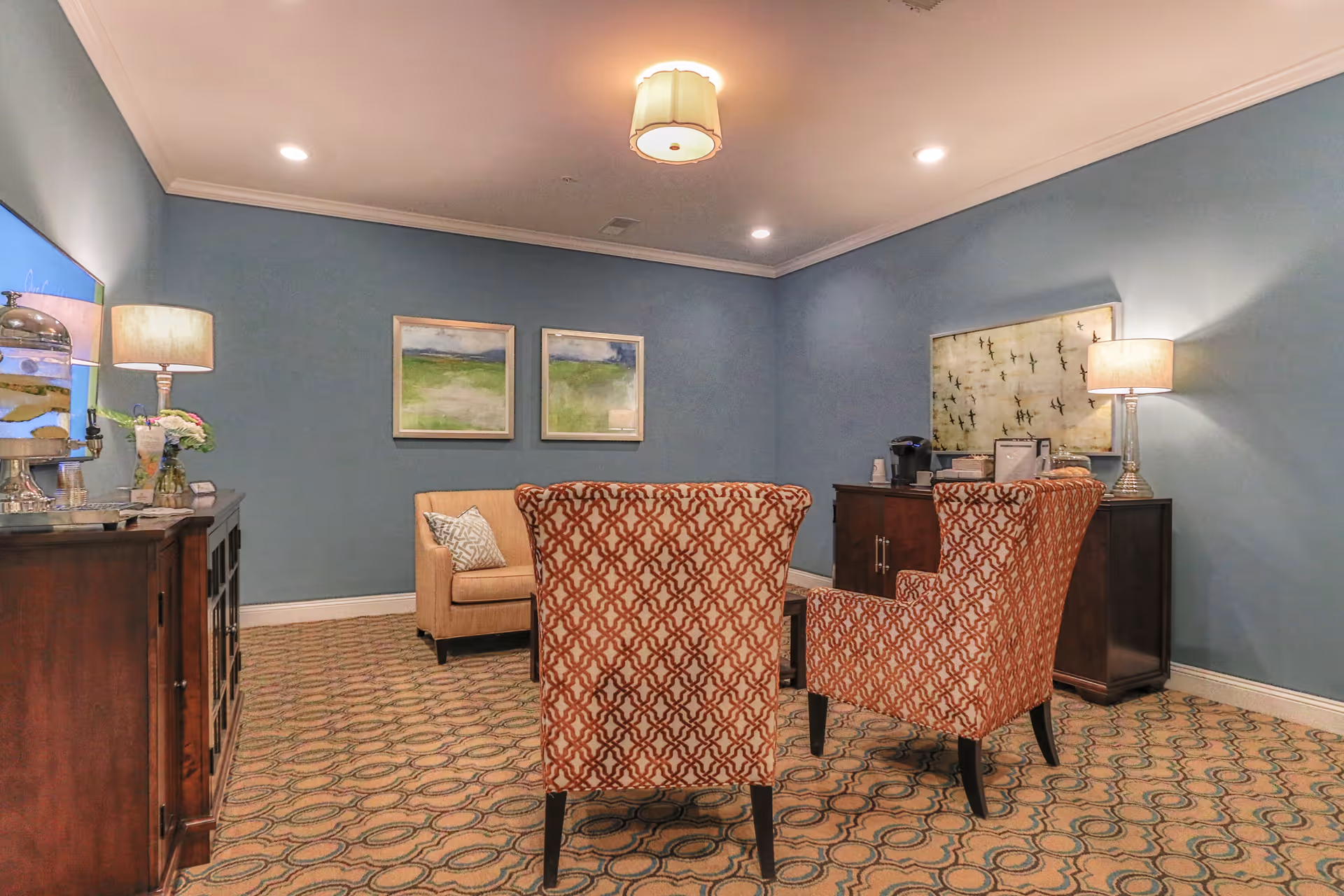 A cozy sitting area with two patterned armchairs facing a beige loveseat against a blue wall. The room features a patterned carpet, two framed landscape paintings on the wall, two table lamps on wooden cabinets, and a coffee station with a coffee maker and supplies.
