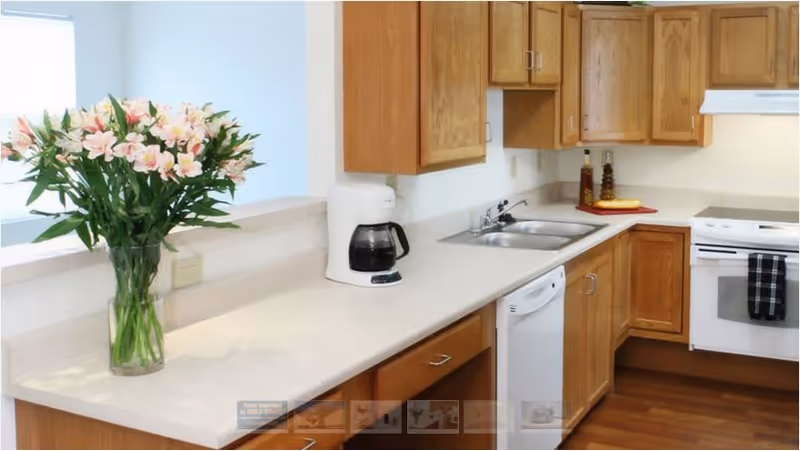 A clean kitchen with wooden cabinets, a white countertop, a coffee maker, a double sink, a dishwasher, and a white stove with a black and white checkered towel hanging on the handle. A vase with pink flowers is placed on the countertop near a window.