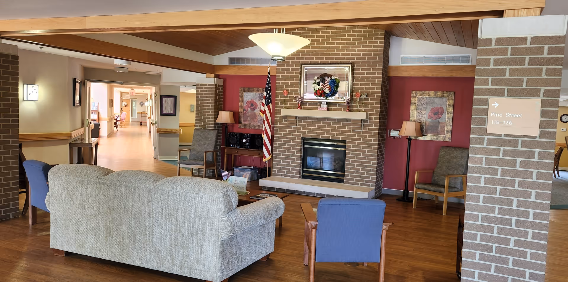 A cozy common area in a senior living facility featuring a beige couch and a blue armchair facing a brick fireplace with an American flag beside it. The room has wooden floors, a wooden ceiling with exposed beams, and red accent walls with framed floral artwork. There are additional chairs and lamps around the room, and a hallway is visible in the background with more seating and natural light.