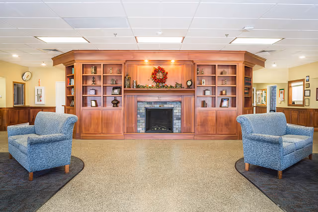 A cozy common area with two blue upholstered armchairs facing a wooden fireplace with built-in shelves. The shelves contain decorative items and books, and a festive wreath hangs above the fireplace. The room has a neutral color scheme with beige walls and a speckled floor.