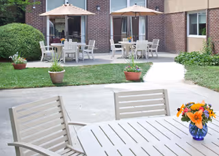 Outdoor patio area with white tables and chairs, some tables shaded by beige umbrellas. Potted plants and flowers are placed around the patio, with a brick building in the background.
