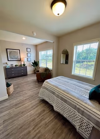 A bright bedroom with a bed covered in a patterned blanket, two large windows with white blinds, a wall-mounted mirror, a brown armchair with a green throw, a dark wooden dresser with decorative items, framed artwork on the wall, and wood-look flooring.
