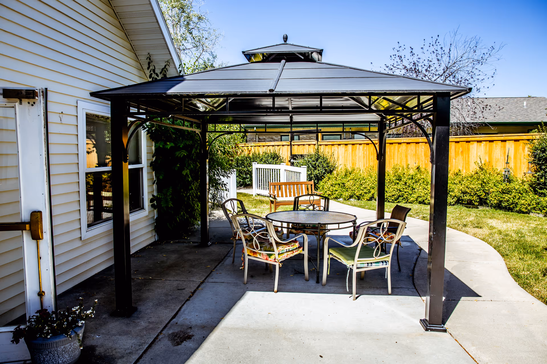 Outdoor patio area with a metal gazebo covering a round glass table surrounded by four chairs with cushions. There is a wooden bench in the background, a concrete pathway, green bushes, and a wooden fence under a clear blue sky.