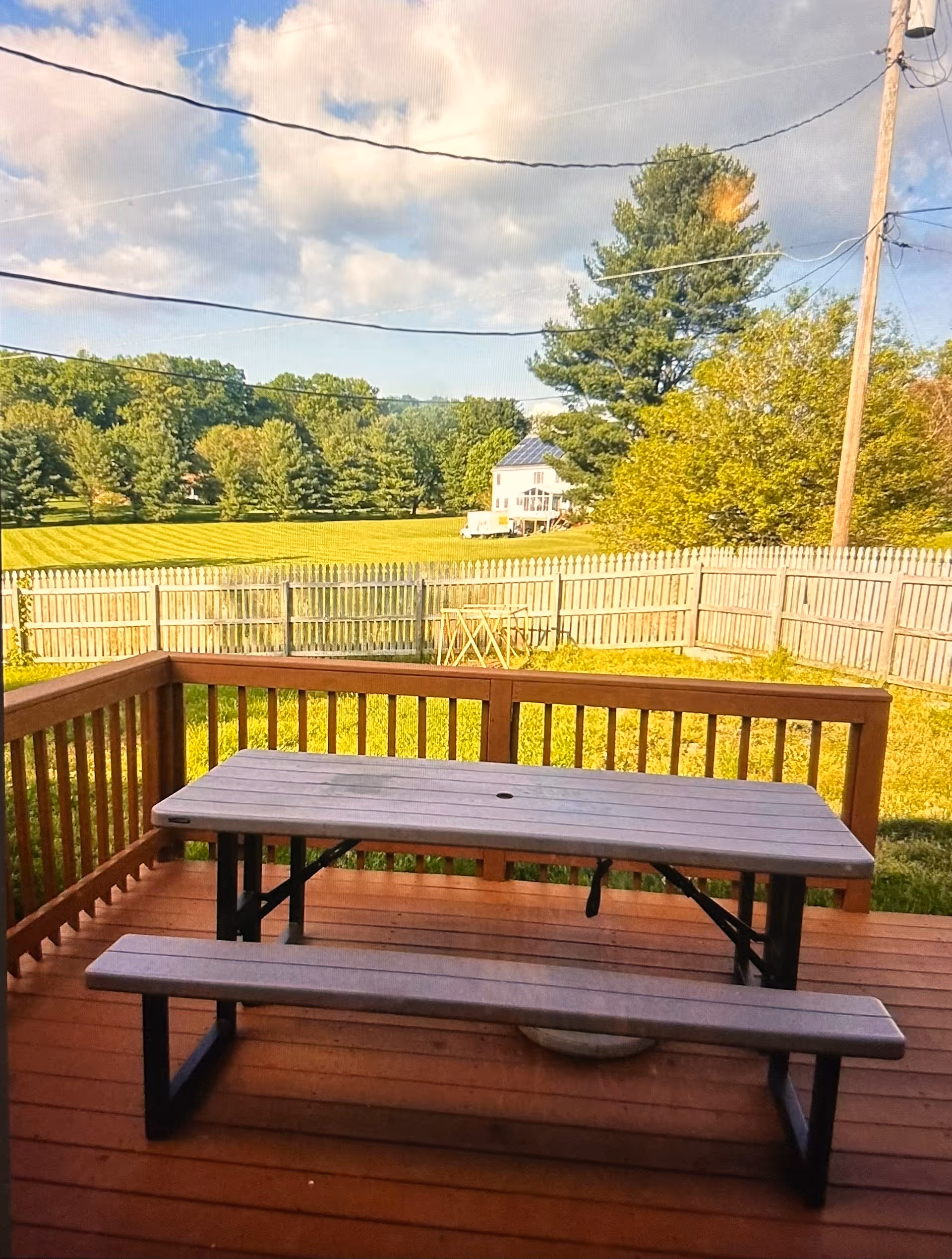A wooden deck with a picnic table and bench overlooking a fenced backyard with green grass and trees. In the background, there is a house with solar panels on the roof under a partly cloudy sky.