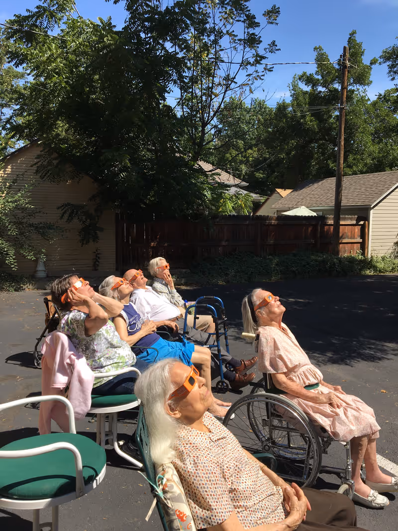 A group of elderly people sitting outdoors in chairs and wheelchairs, all wearing protective eclipse glasses and looking up at the sky. They are in a paved area with trees, a wooden fence, and houses in the background under a clear blue sky.