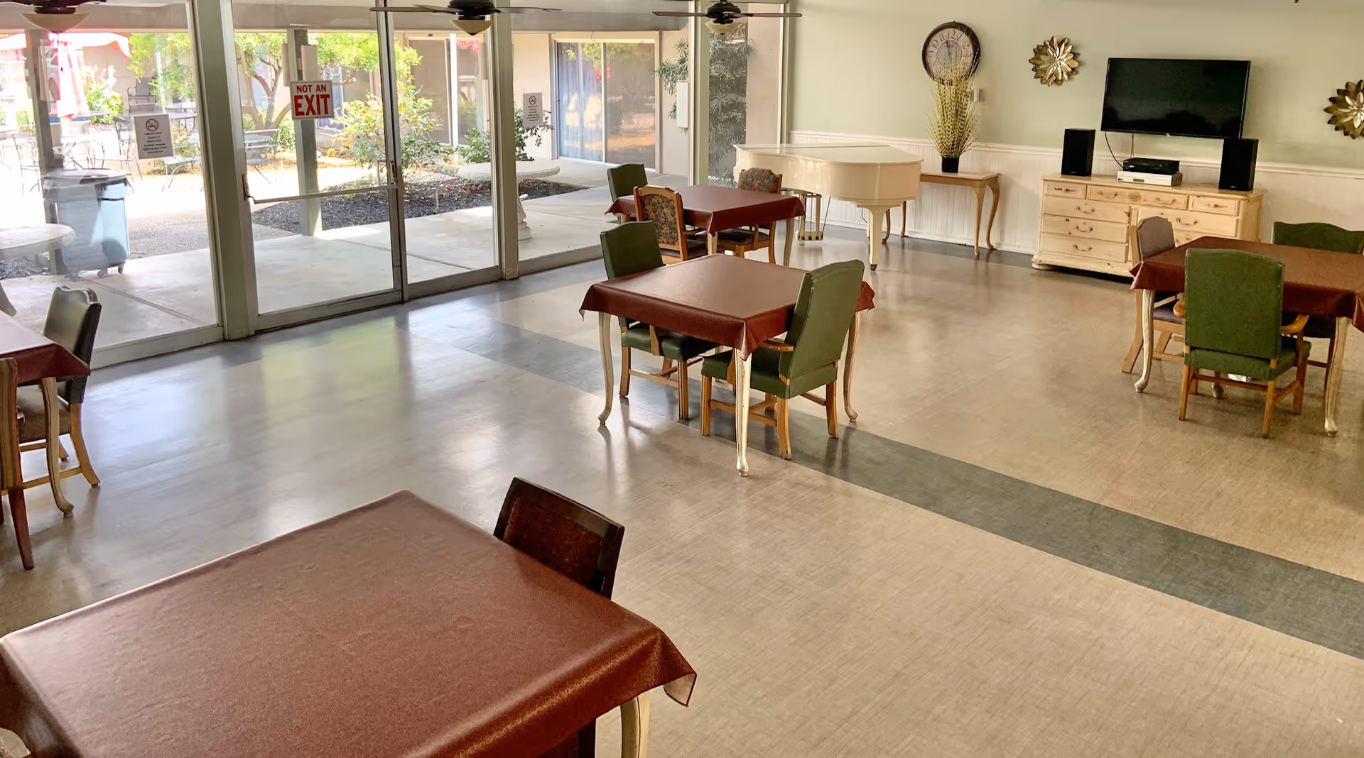 A spacious common area with several tables covered in brown tablecloths and green chairs around them. Large glass sliding doors lead to an outdoor patio area with trees and seating. Inside, there is a white piano, a TV mounted on the wall above a wooden dresser, and decorative wall hangings. The room has a light-colored floor with a darker strip running through the middle.