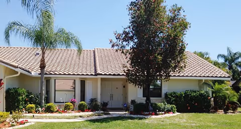 Single-story residential building with a tiled roof, a front door in the center, windows on either side, surrounded by palm trees, bushes, and a well-maintained lawn under a clear blue sky.