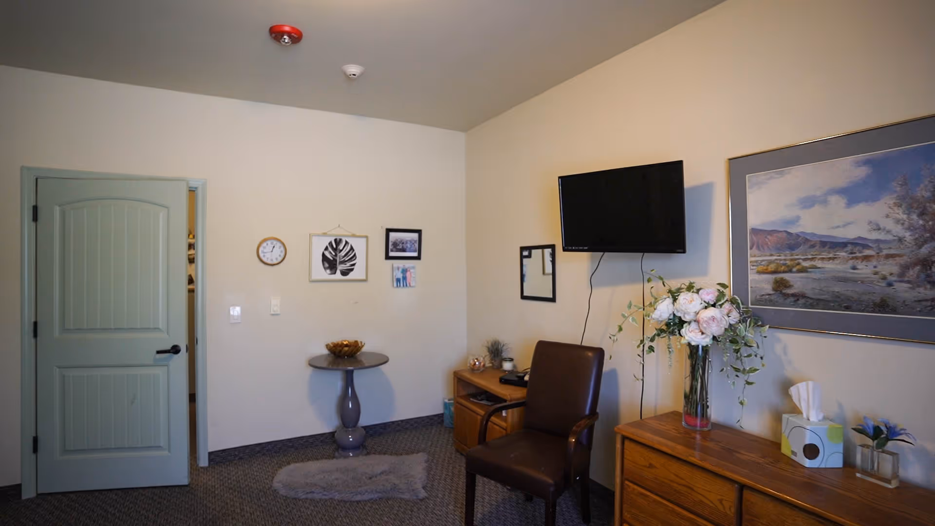 Interior room with beige walls and carpeted floor featuring a green door, a small round table with a decorative bowl, a brown chair, a wooden dresser with a vase of flowers and a tissue box, a wall-mounted TV, and framed pictures on the walls.