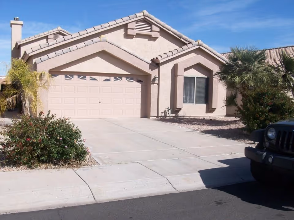 Front exterior view of a single-story house with a two-car garage, beige stucco walls, and a tiled roof. There are some bushes and palm trees in the front yard, and a black vehicle is partially visible on the right side of the driveway under a clear blue sky.