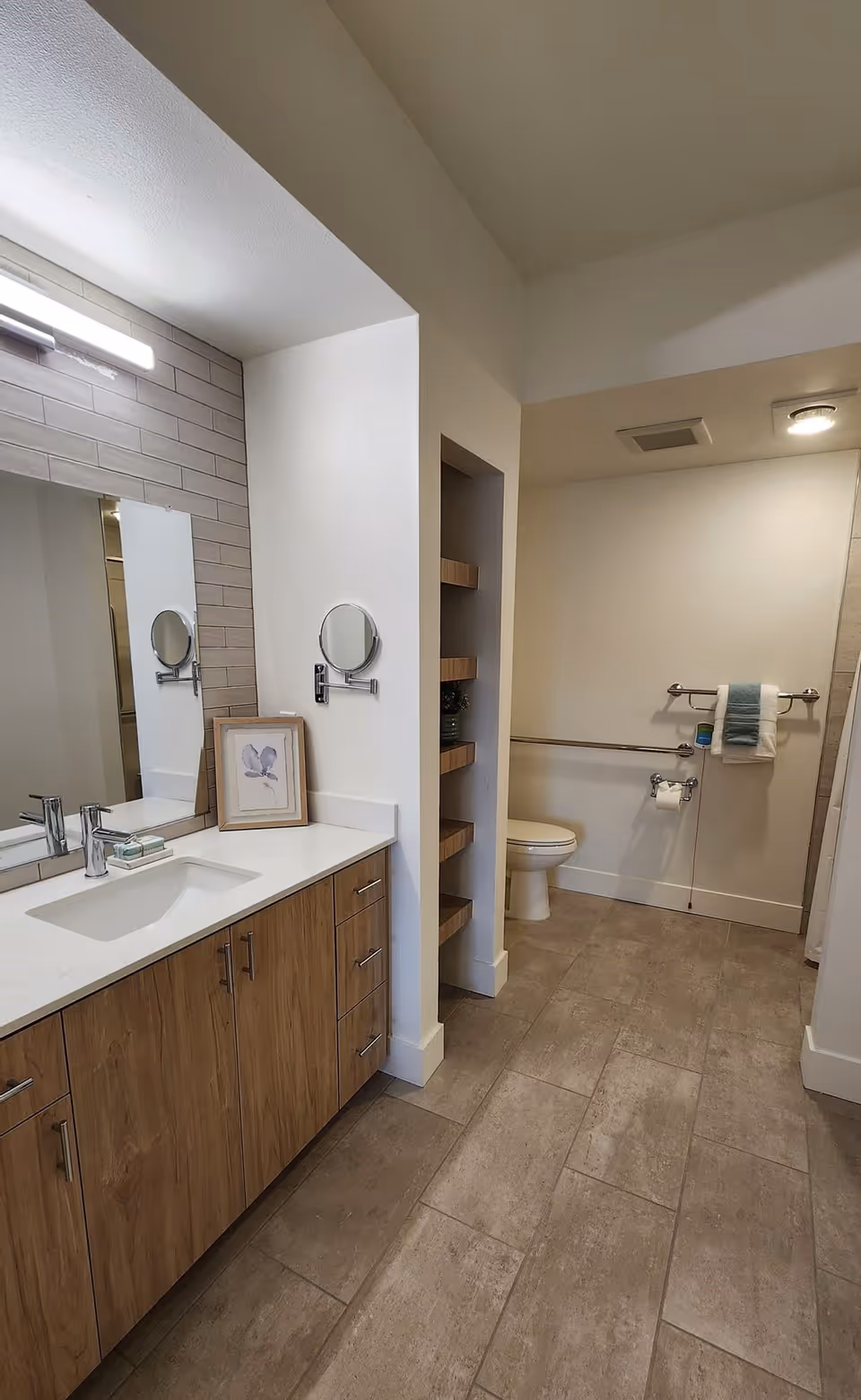 A modern bathroom with a wooden vanity featuring a white countertop and a rectangular sink. Above the vanity is a large mirror with a light fixture. On the countertop, there is a framed picture and a soap dish. To the right, there is a toilet with grab bars on the wall and a towel hanging on one of the bars. The floor is tiled in a neutral color, and there are built-in wooden shelves next to the toilet.