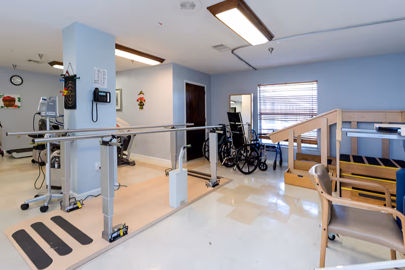 Rehabilitation room with parallel bars for walking exercises, a wheelchair, a walker, exercise equipment, a wooden ramp with handrails, and a chair. The room has light blue walls, a window with blinds, and overhead fluorescent lighting.