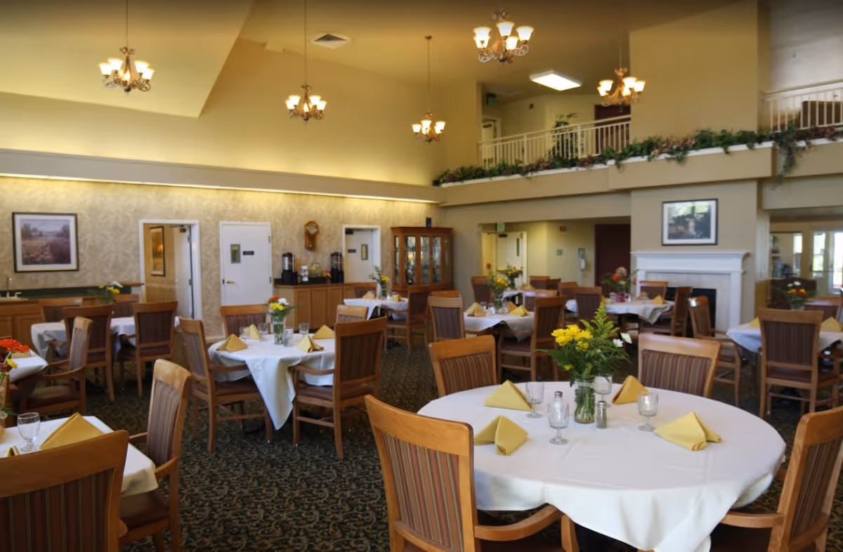 A spacious dining room with round tables covered in white tablecloths, each set with yellow folded napkins, glassware, and floral centerpieces. The room features wooden chairs with striped cushions, a patterned carpet, chandeliers hanging from a high ceiling, and a fireplace with a framed picture above it. There is a balcony with plants and railings overlooking the dining area.