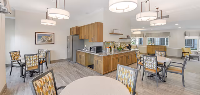 A bright and spacious dining area with multiple round tables and patterned chairs. The room features wood cabinetry with a granite countertop, a stainless steel refrigerator, a microwave, and beverage dispensers. Large windows allow natural light to fill the space, and modern pendant lights hang from the ceiling.