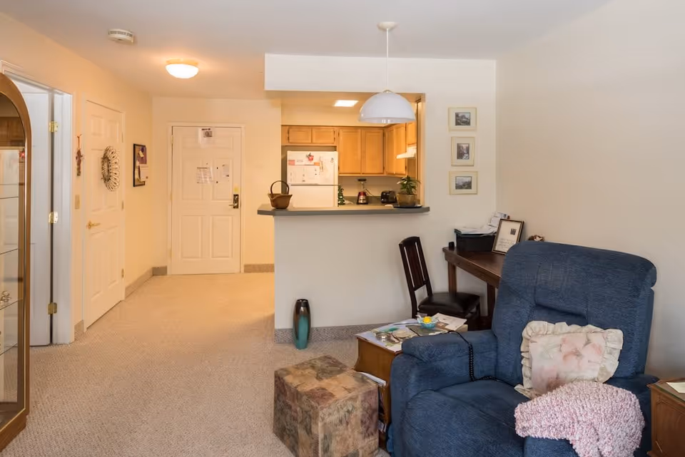 Interior view of a senior living facility apartment showing a blue recliner chair with a floral pillow and pink knitted blanket, a small wooden table with magazines, a patterned ottoman, a wooden desk with a chair, and a kitchen area with light wood cabinets and a white refrigerator in the background.
