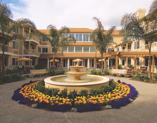 Outdoor courtyard of a senior living facility with a central tiered fountain surrounded by colorful flowers and palm trees. The courtyard is enclosed by a multi-story building with balconies and large windows. Benches and shaded seating areas are visible around the courtyard.