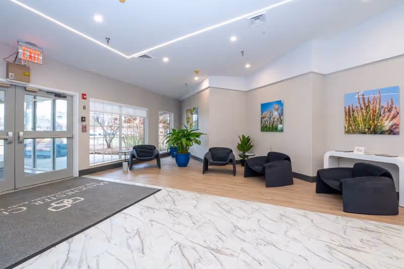 A bright and modern facility lobby area with a marble floor near the entrance and wood flooring in the seating area. The seating area has five black chairs arranged around two small tables, with two large potted plants. The walls are light-colored and decorated with colorful nature-themed artwork. Large windows allow natural light to enter, and there is an exit sign above the glass doors.