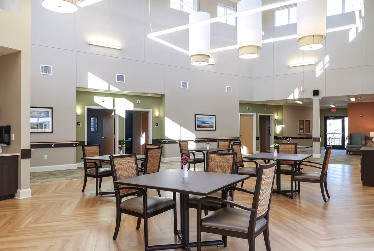 A bright and spacious common area in a senior living facility with several square tables and chairs arranged neatly. The room features high ceilings with large windows allowing natural light to fill the space. There are framed pictures on the walls and small flower vases on each table. The flooring is wood-patterned, and the walls are painted in neutral tones with some green accents around doorways.