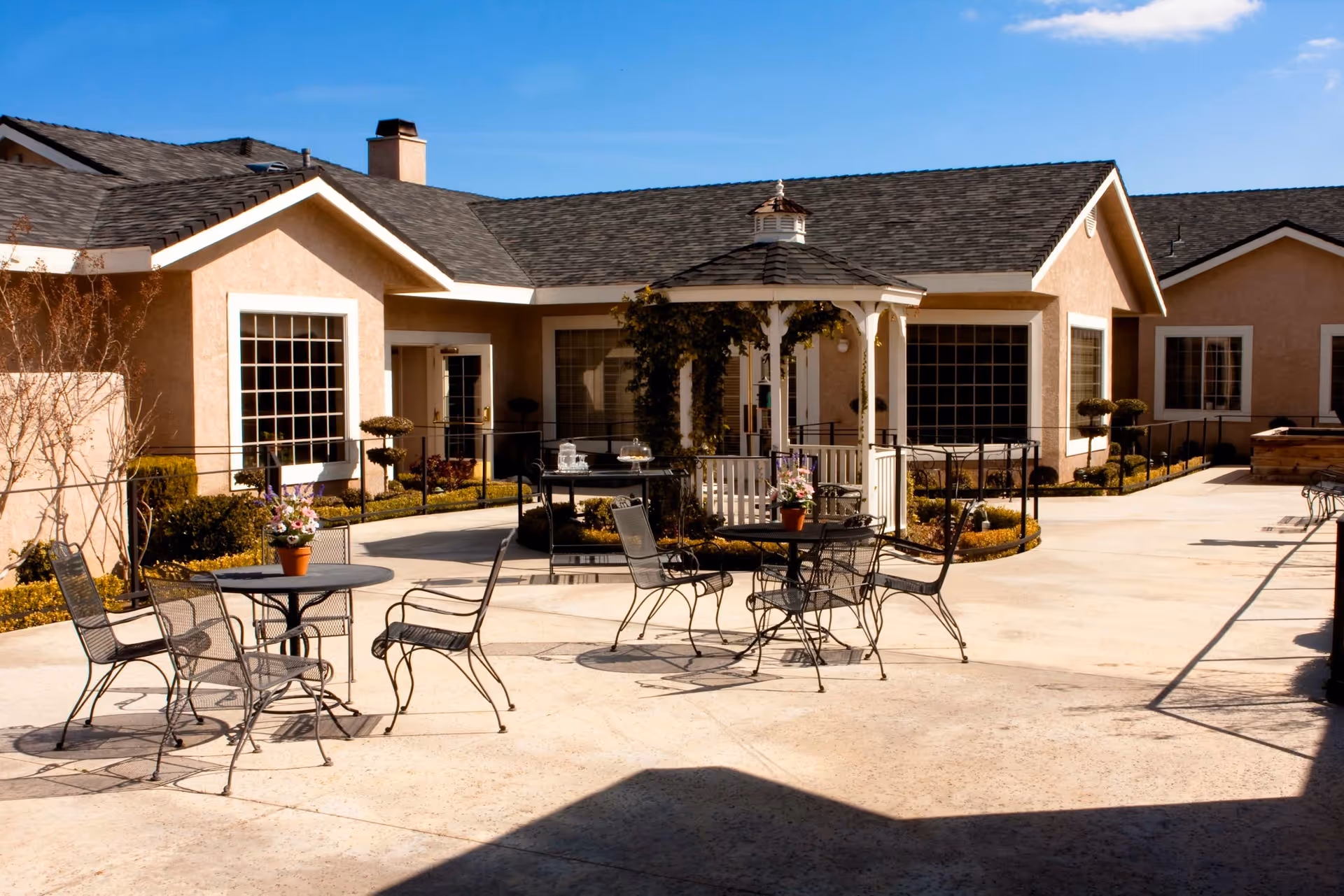 Outdoor courtyard area of Sterling Commons Memory Care Community featuring metal tables and chairs with potted flowers, a white gazebo with greenery, and beige buildings with large windows under a clear blue sky.