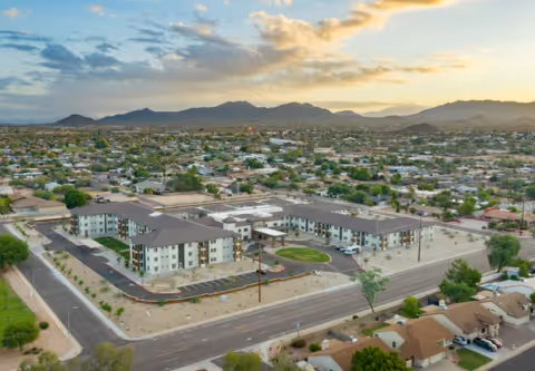 Aerial view of a three-story U-shaped senior living complex with parking, surrounding neighborhood, and distant mountains at sunset.
