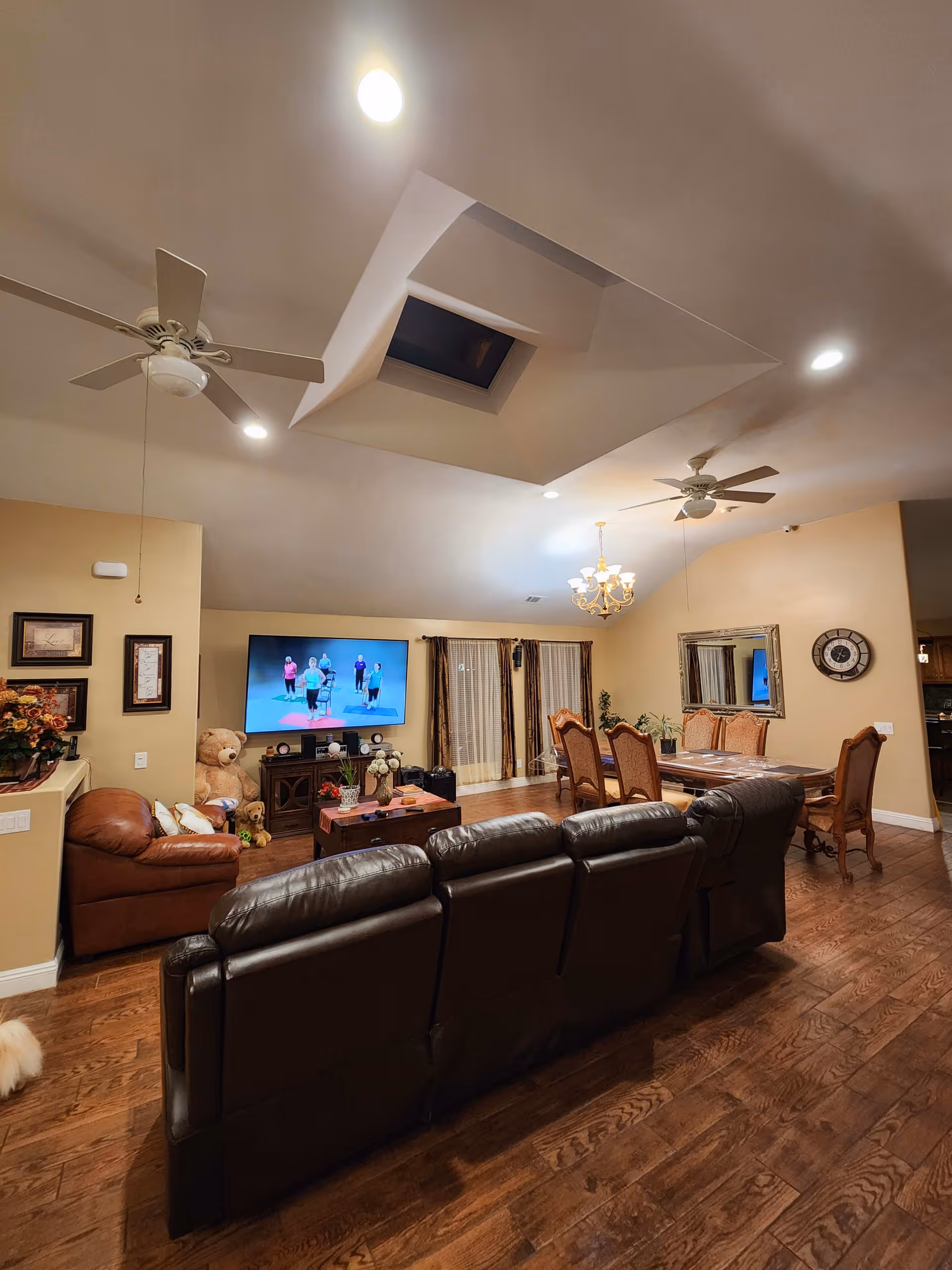 A spacious living room and dining area with wooden flooring, beige walls, and a vaulted ceiling with two ceiling fans and a skylight. The living room has a large dark leather sofa facing a mounted flat-screen TV displaying a fitness video. There is a brown leather armchair with a large teddy bear beside it. The dining area features a wooden dining table with six chairs, a chandelier, a large wall mirror, and a decorative wall clock. French doors with curtains lead outside.