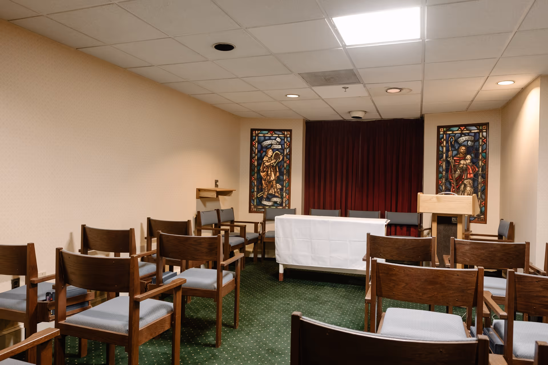 A small chapel or meeting room with rows of wooden chairs with gray cushions facing a table covered with a white cloth and a wooden podium. The room has green carpet, beige walls, a drop ceiling with fluorescent lighting, and two stained glass windows depicting religious figures on the back wall.