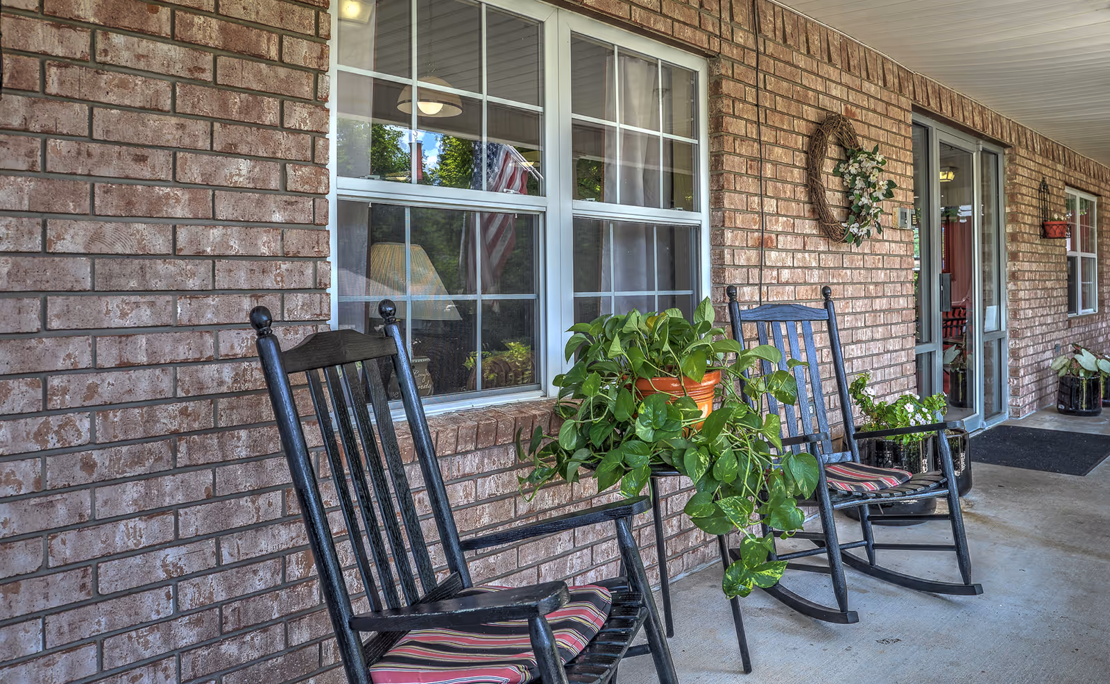 A brick front porch with two black rocking chairs, potted plants, and windows.