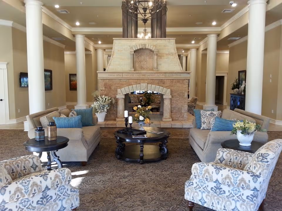 Elegant common lounge with sofas and armchairs arranged around a central stone fireplace, decorative columns, and a chandelier.
