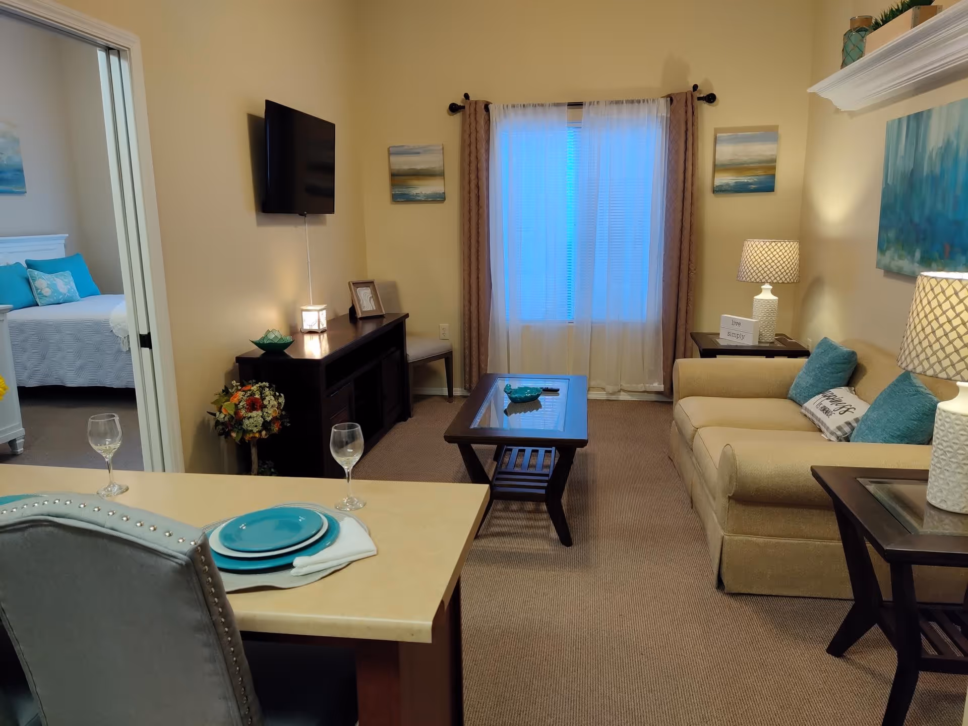 A cozy living room area with a beige sofa adorned with blue and white pillows, two side tables with lamps, a coffee table, and a TV mounted on the wall. The room has a window with sheer curtains and brown drapes. In the foreground, there is a dining counter with a place setting including a plate, napkin, and wine glass. A bedroom with a bed and blue pillows is visible through an open doorway.