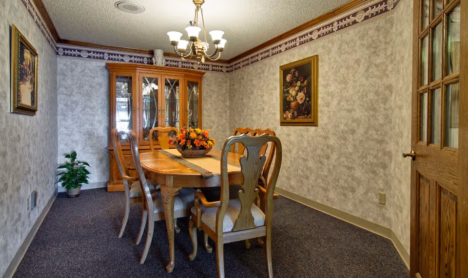 A traditional dining room with a wooden dining table and six matching chairs. The table has a floral centerpiece and a table runner. There is a wooden china cabinet with glass doors against the back wall. The walls are decorated with patterned wallpaper and framed floral paintings. A wooden door with glass panels is partially visible on the right side, and a small potted plant is on the left side near the wall.