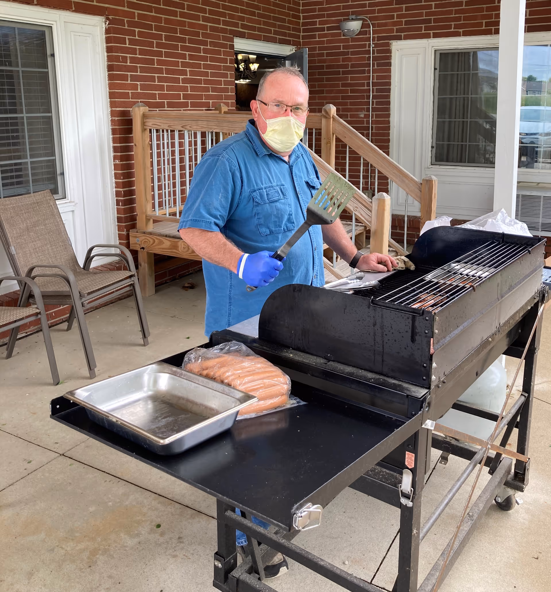 A man wearing a blue shirt, yellow face mask, and blue gloves is standing outdoors next to a large black grill. He is holding a spatula and appears to be grilling food. There is a package of sausages and a metal tray on the grill's side table. Behind him is a brick wall, a wooden ramp with railings, and patio chairs.