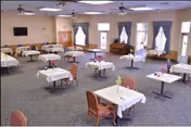 A spacious dining room with multiple tables covered in white tablecloths, each set with a small vase of flowers. The room has several windows with curtains, a television mounted on the wall, and a piano in the corner. Chairs are arranged around the tables, and the carpeted floor is visible.