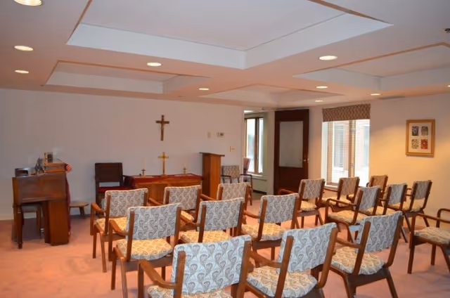 A small chapel or prayer room with rows of wooden chairs upholstered in patterned fabric facing a small altar with a cross on the wall behind it. There is a piano on the left side and a door and window on the right side of the room.