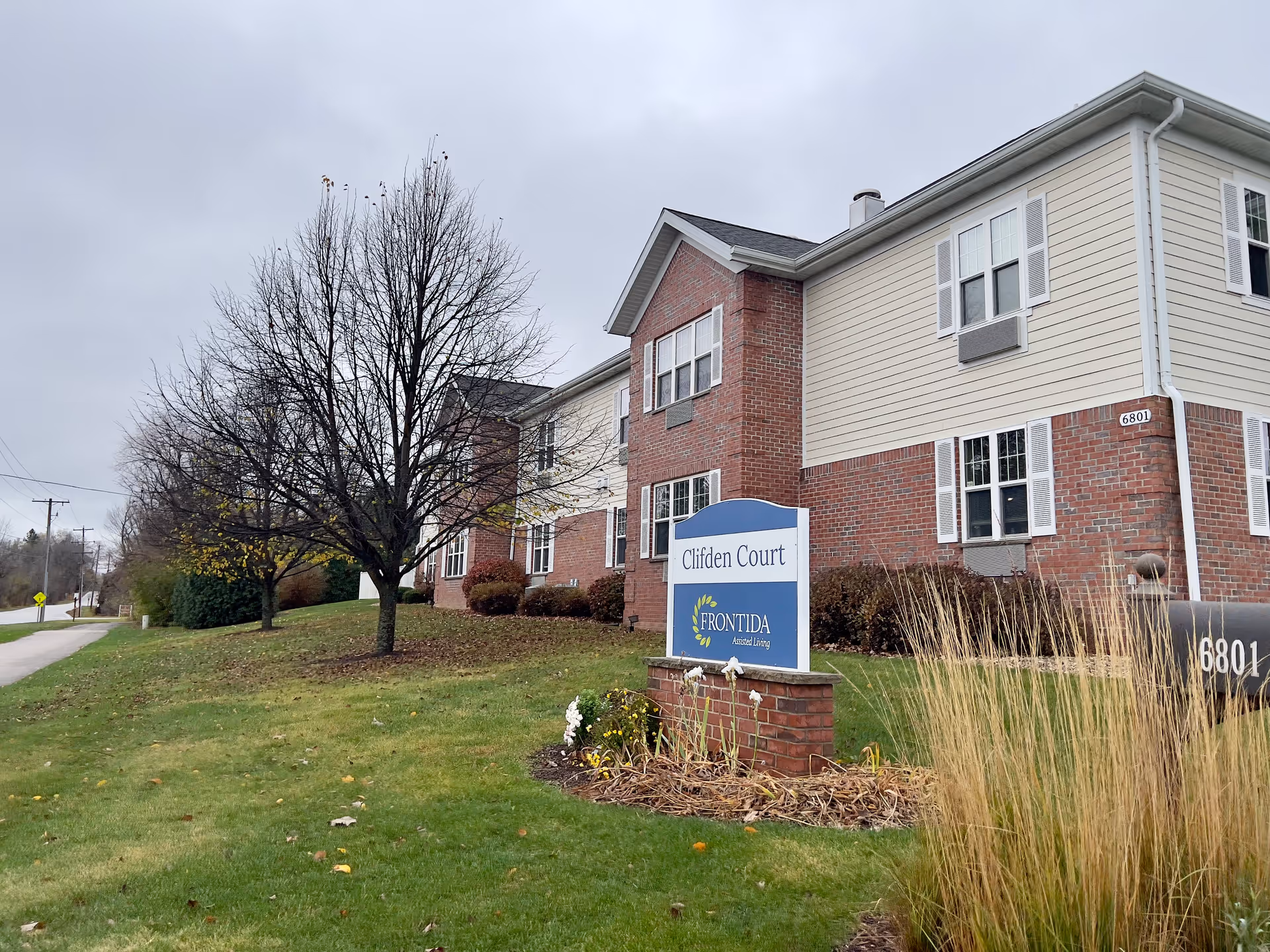 Exterior view of a two-story assisted living facility named Clifden Court with a sign in front that reads 'Clifden Court Frontida Assisted Living'. The building has brick and beige siding with multiple windows. There is a grassy area with some trees and shrubs, and a cloudy sky overhead.