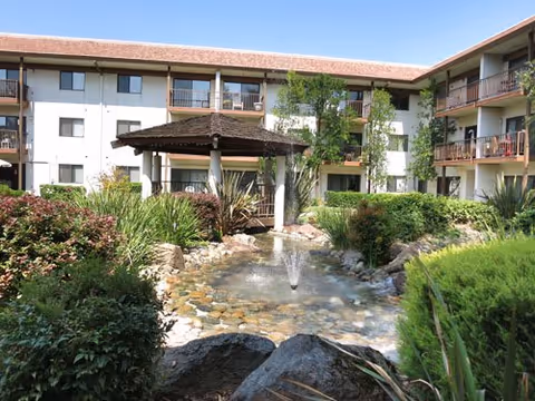 Outdoor courtyard area at Roseville Commons Senior Living featuring a central water fountain surrounded by rocks and lush greenery, with a wooden gazebo and a three-story building with balconies in the background under a clear blue sky.