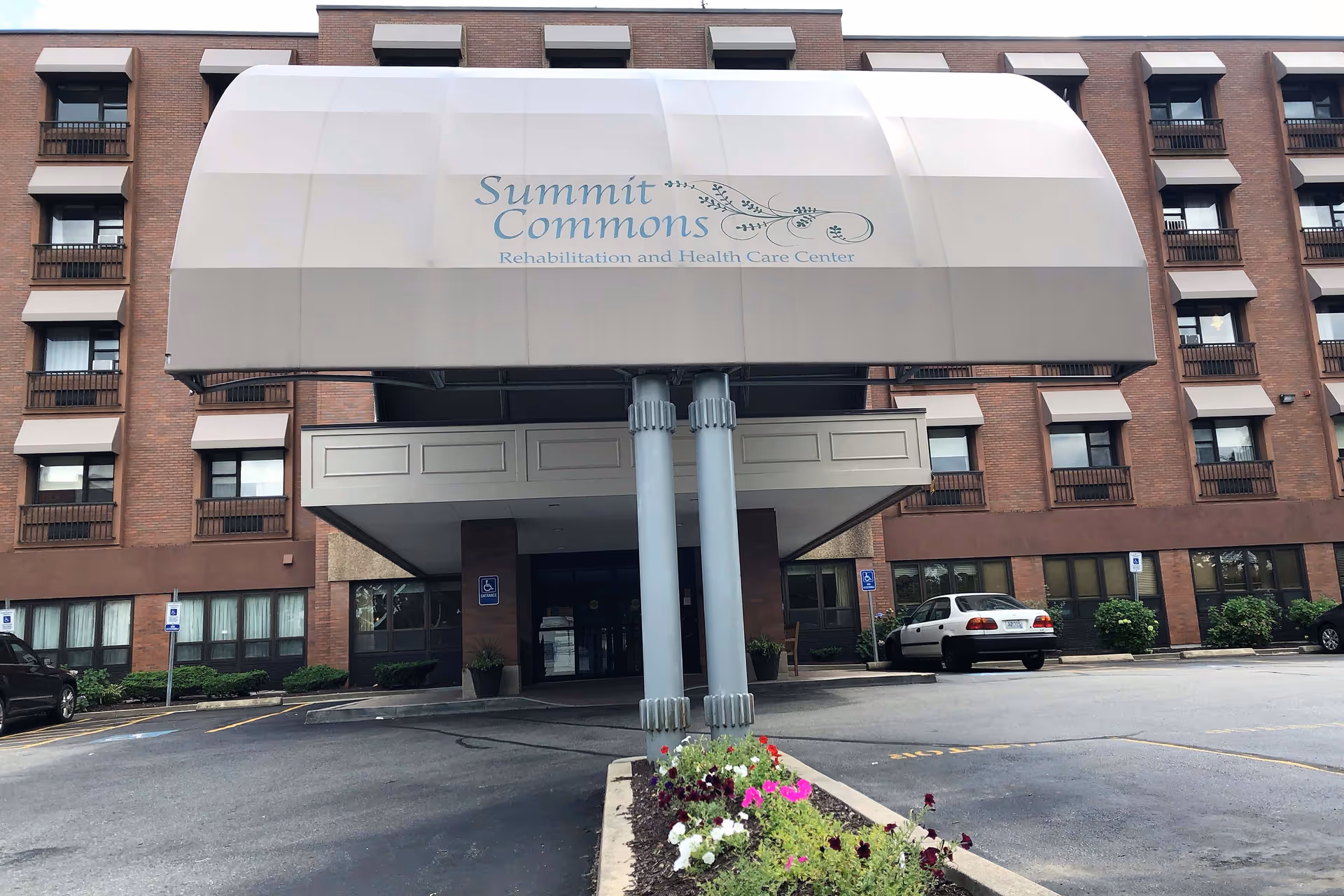 Front entrance of Summit Commons Rehabilitation and Health Care Center, showing a large beige awning with the facility's name and logo. The building is a multi-story brick structure with several windows and small balconies. There are a few parked cars and a flower bed with colorful flowers in front of the entrance.