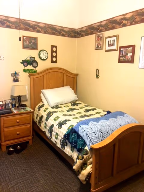 A small bedroom with a single wooden bed featuring a tractor-themed blanket and a blue knitted throw. Next to the bed is a wooden nightstand with a lamp, framed photos, and other small items. The walls are decorated with framed pictures, a clock, and a tractor wall decoration. The room has a carpeted floor and a wallpaper border near the ceiling with a nature or animal theme.