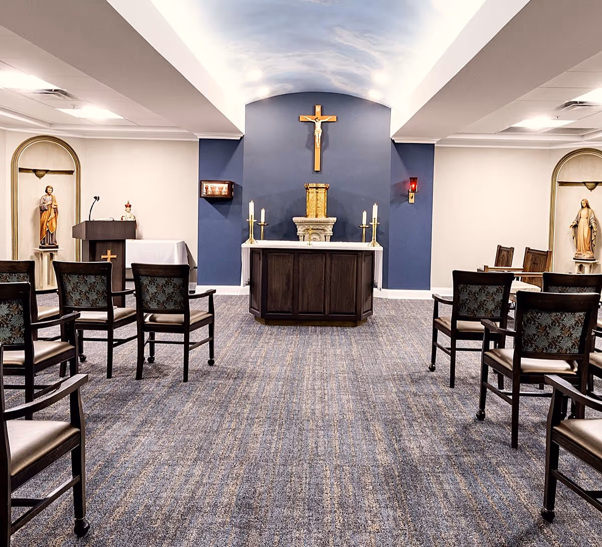 Interior view of a chapel room with rows of chairs facing an altar. The altar has candles and a crucifix mounted on a blue accent wall. Statues are placed in alcoves on either side of the room, and a lectern with a microphone is visible on the left side.