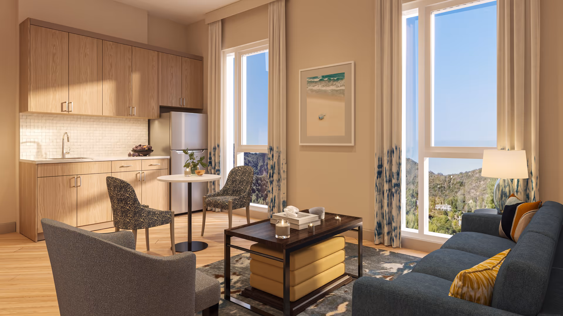 A bright and modern senior living apartment interior featuring a small kitchen area with wooden cabinets, a round dining table with two patterned chairs, a coffee table with candles and books, a blue sofa with yellow and patterned cushions, large windows with curtains offering a scenic view of hills and greenery outside.