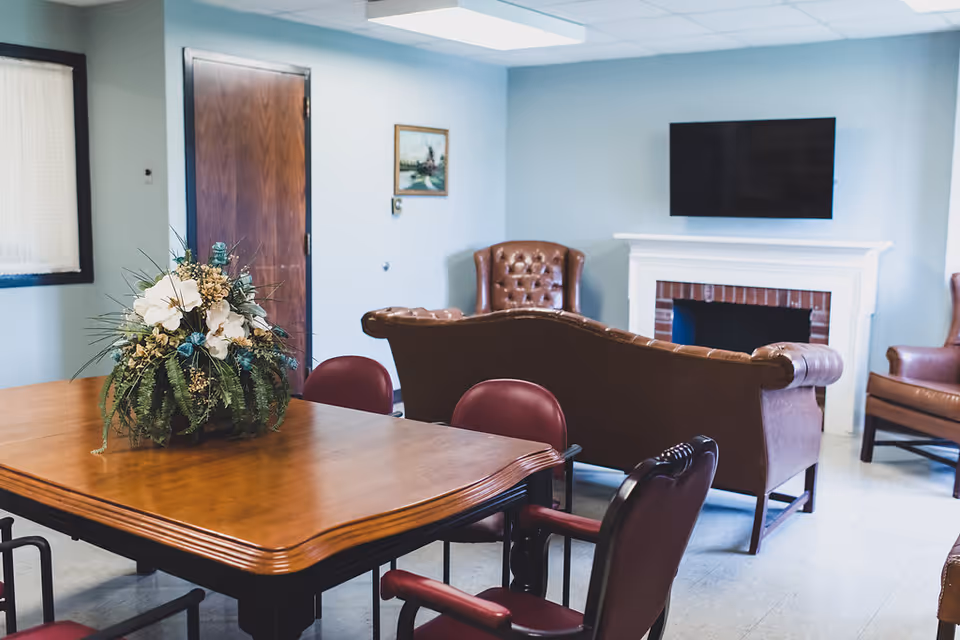 A cozy sitting area in a nursing center featuring a wooden table with a floral arrangement, surrounded by maroon chairs. In the background, there is a brown leather sofa and armchairs facing a white fireplace with a mounted flat-screen TV above it. The walls are painted light blue, and there is a wooden door and a framed painting on the wall.