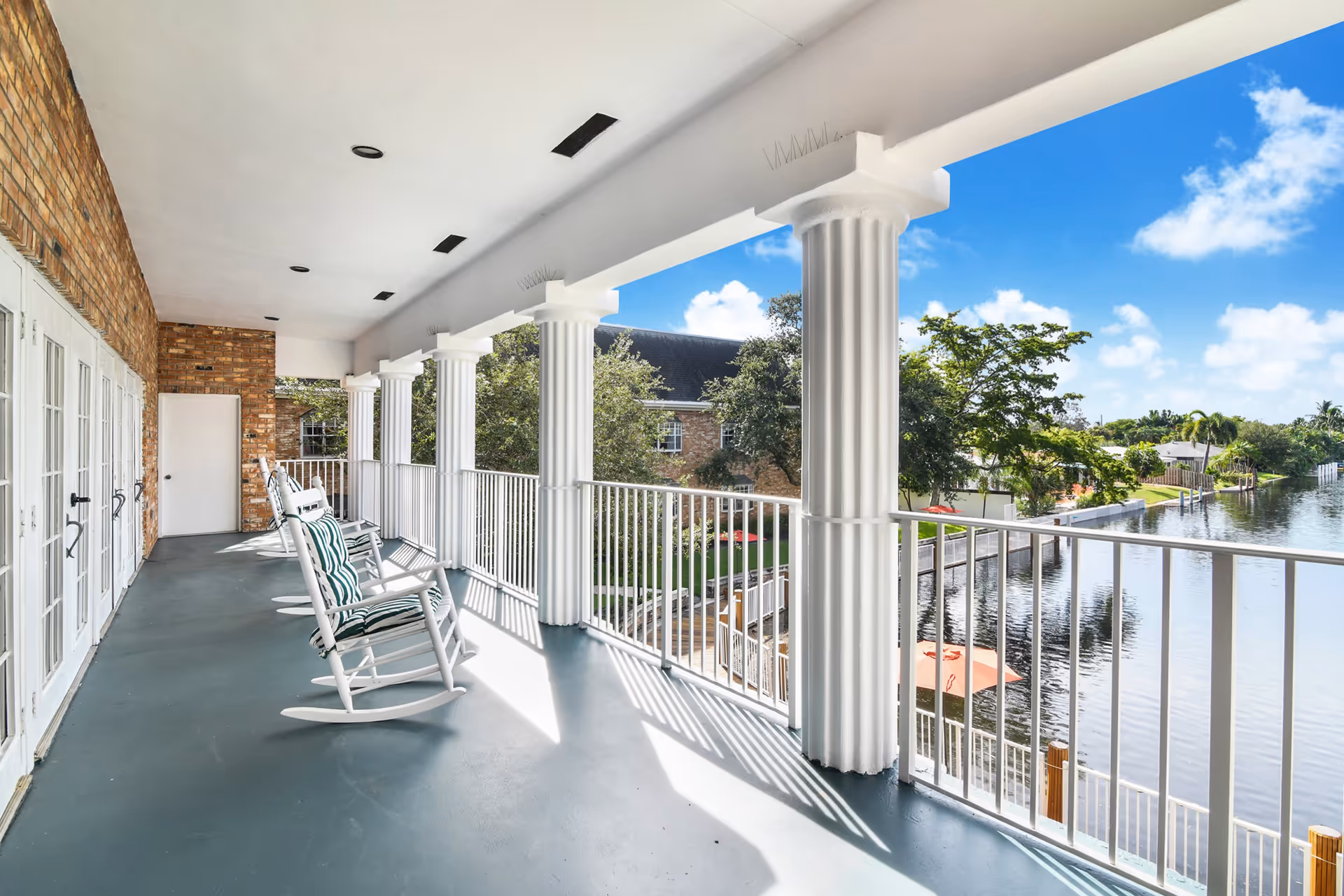A spacious covered balcony with white rocking chairs featuring striped cushions, white railings, and large white columns overlooking a calm waterway with trees and houses on the opposite shore under a blue sky with scattered clouds.
