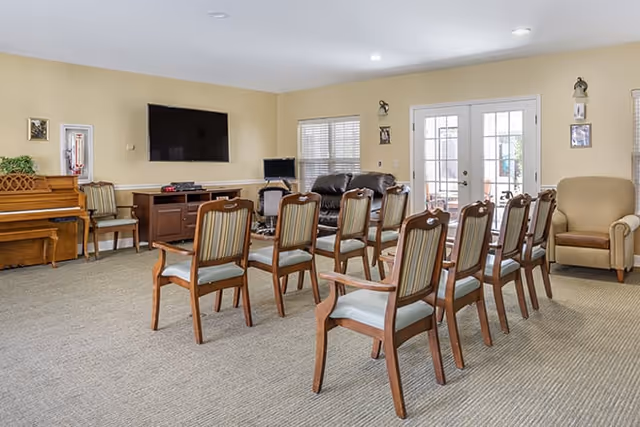 Communal living room with rows of wooden chairs facing a wall-mounted TV, a piano, and French doors.