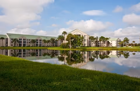 A large senior living facility building with a green roof and multiple windows, reflected in a calm pond in front of it. The sky is partly cloudy and there is green grass surrounding the pond.