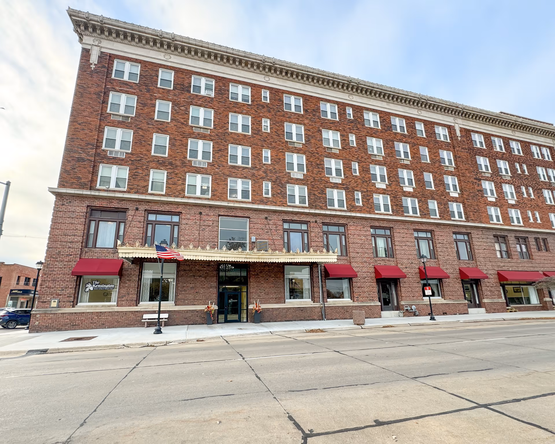Exterior view of a large multi-story brick building with numerous windows and red awnings over the ground floor windows. The building has a decorative cornice and an entrance canopy with ornamental details. An American flag is mounted on a pole near the entrance, and there is a sign on the left side window that reads 'The Kensington'. The street in front of the building is empty.