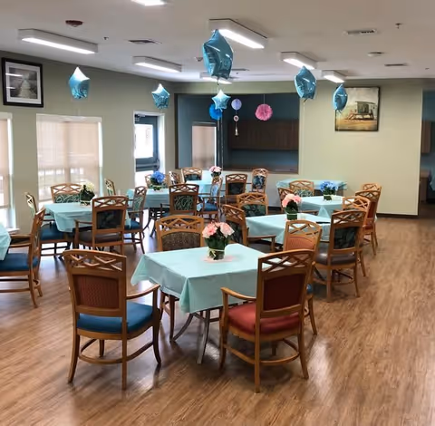 A dining room with multiple tables covered in light blue tablecloths, each adorned with a small flower arrangement. Wooden chairs with blue and red cushions surround the tables. Blue star-shaped balloons are tied to some chairs and float above the tables. The room has wooden flooring, light-colored walls, and several windows with blinds partially drawn. There are framed pictures on the walls and ceiling lights providing illumination.