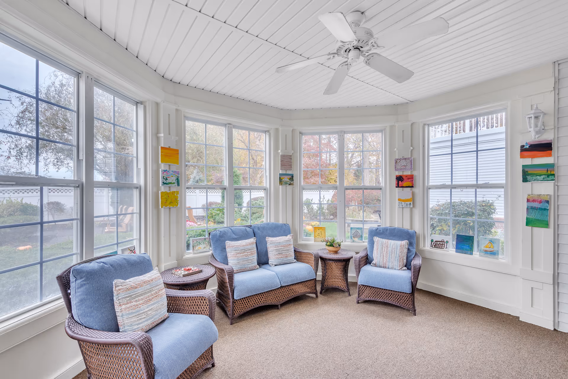 Sunlit enclosed sitting room with wicker armchairs upholstered in blue cushions arranged around a bay of windows under a ceiling fan.