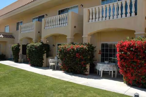 Covered outdoor patio area with white tables, chairs, and red flowering hedges in front of building.