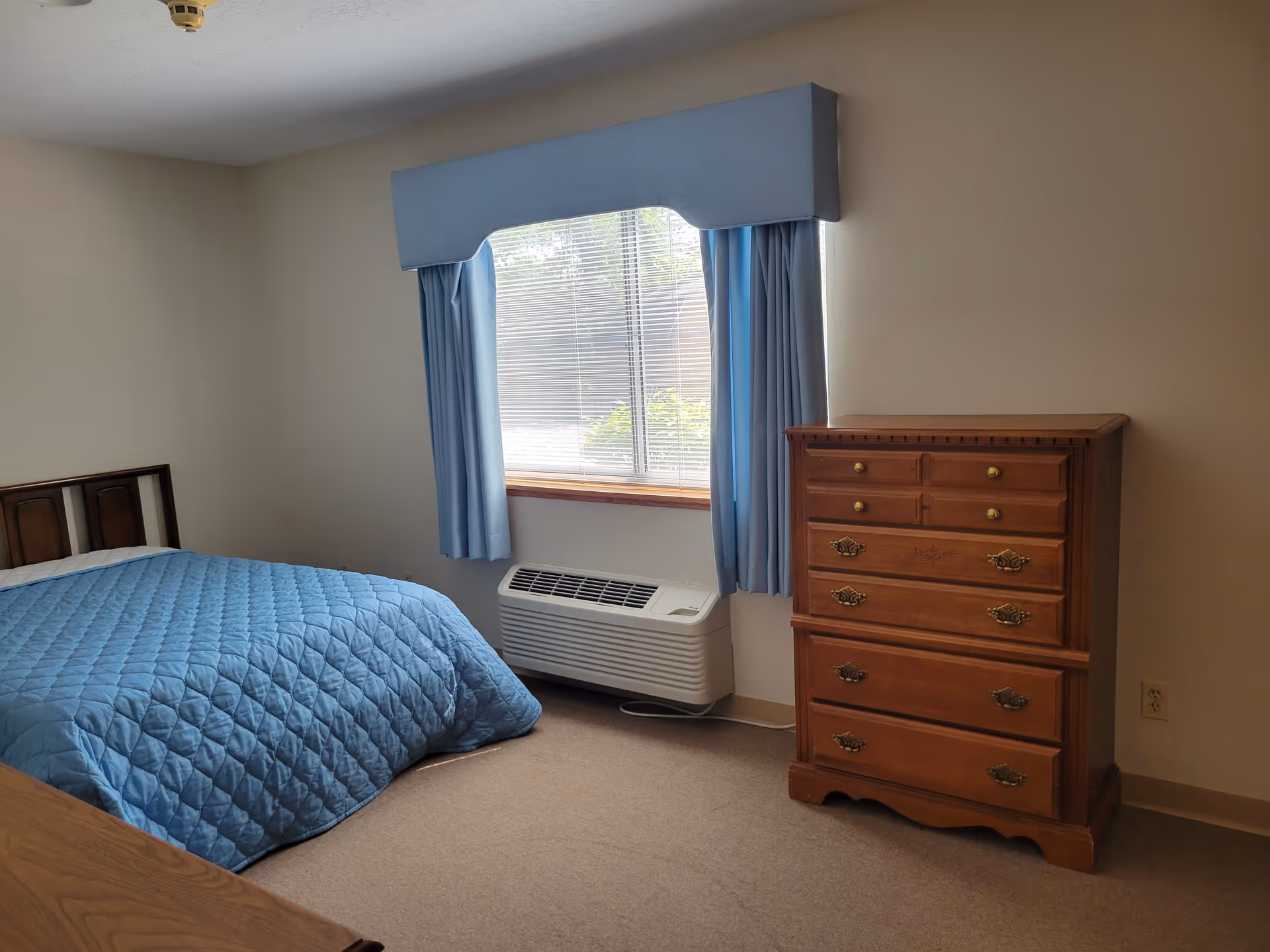 A simple bedroom with a bed covered in a blue quilt, a wooden dresser with multiple drawers, a window with blue curtains and a valance, and a heating/cooling unit beneath the window.
